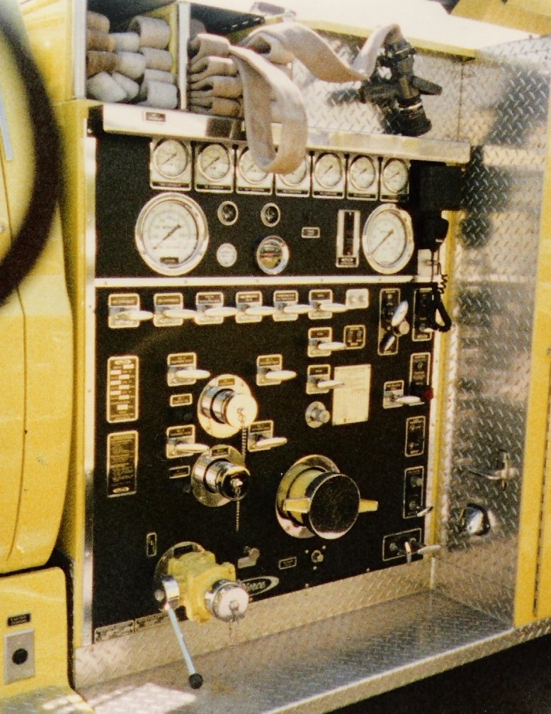 Close-up of a fire truck control panel with various gauges, switches, and dials, including a fire hose nozzle at the bottom left and hoses stored on top.