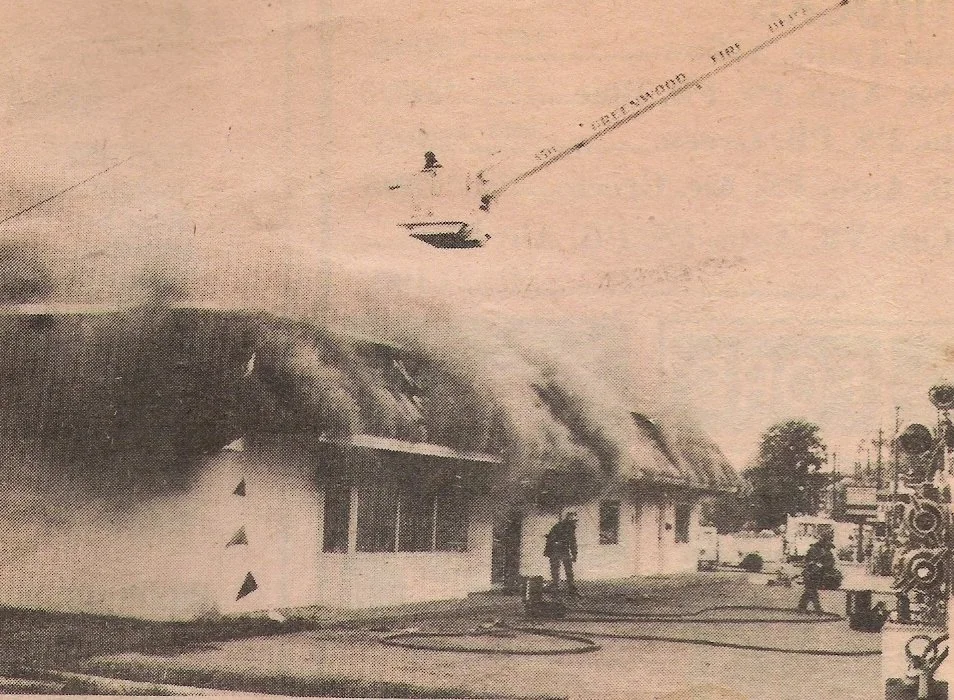 A building on fire with smoke billowing out, and firefighters working to extinguish the blaze. An aircraft is flying overhead.