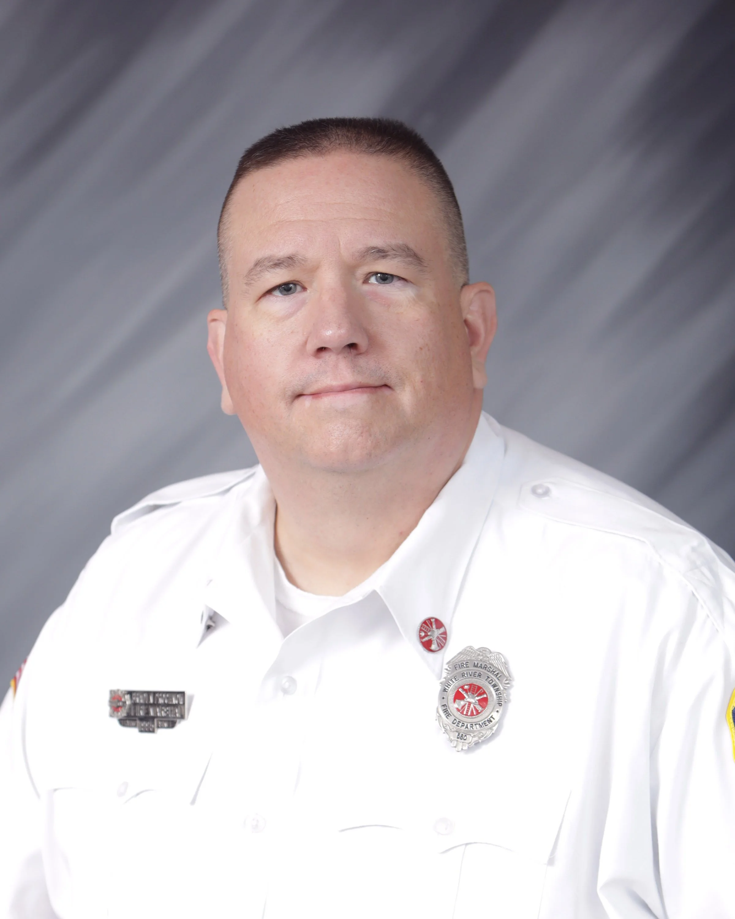 Headshot of a man in a white fire department uniform with badges and insignia, against a gray background.