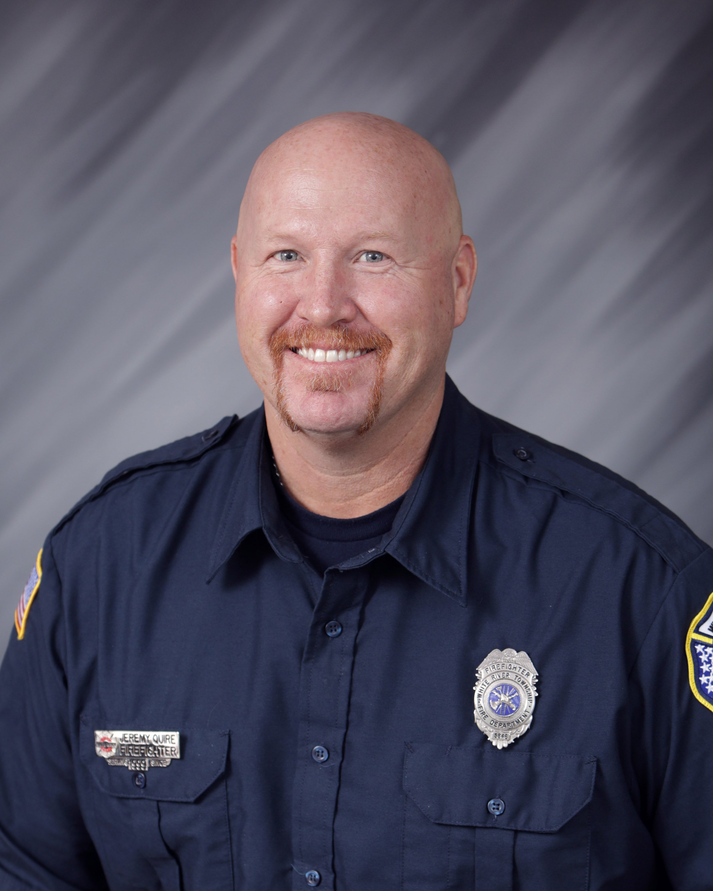 Portrait of a smiling firefighter in uniform with a badge and name tag, against a gray and black streaked background.