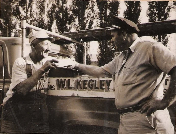 A man in a postal uniform giving a slice of cake to an elderly man wearing a cap and work overalls, with a postal truck in the background.