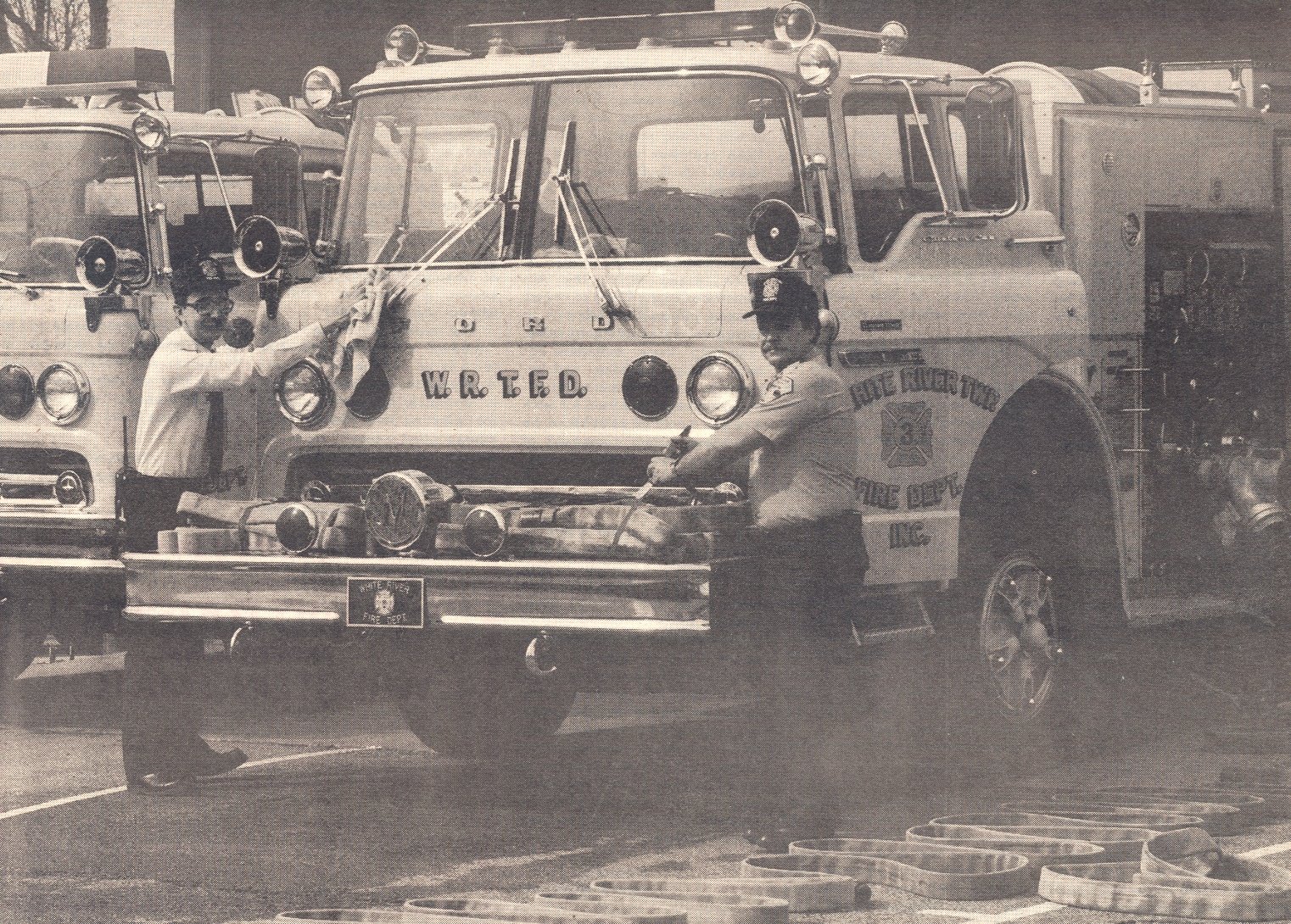 Black and white photo of two firefighters cleaning a fire truck in a parking lot, with other fire trucks in the background.