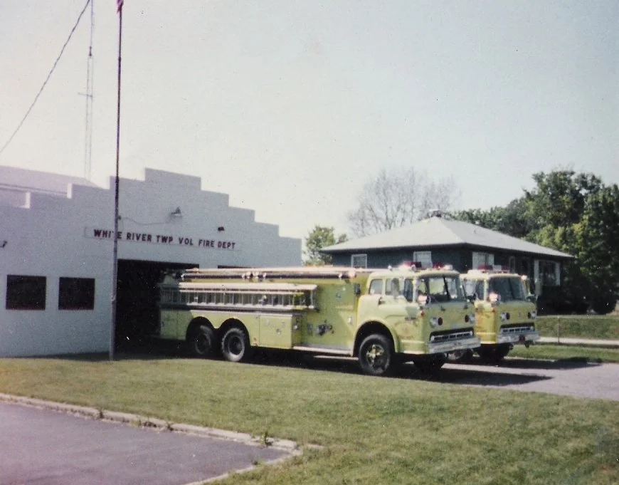 Two vintage yellow fire trucks parked in front of a station building labeled 'White River Twp Vol Fire Dept' with a house and trees in the background.