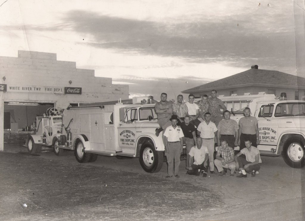 Group of people posing with fire trucks outside a fire station in black and white photo.