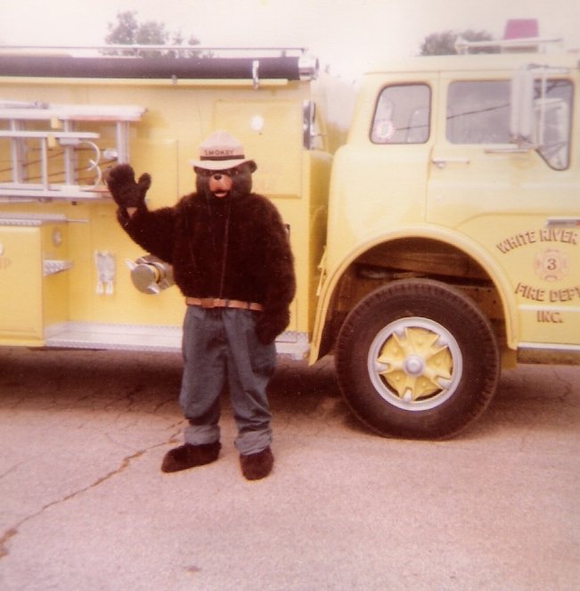 Child dressed in a bear mascot costume standing next to a yellow fire truck, waving with one hand.