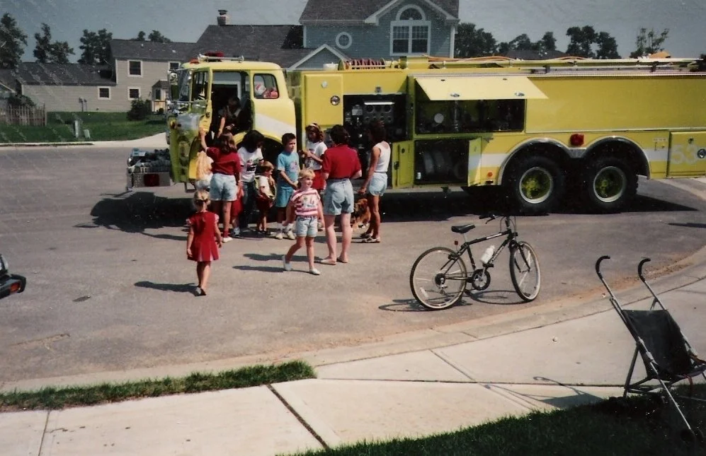 Children and adults gather around a yellow fire truck in a neighborhood, with houses and parked cars in the background. A bicycle and a stroller are on the sidewalk.