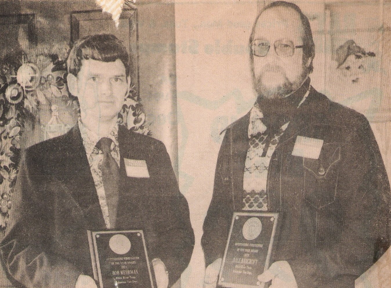 A black-and-white photo of a woman and a man holding plaques or awards, wearing name tags, in an indoor setting with some decorative background items.