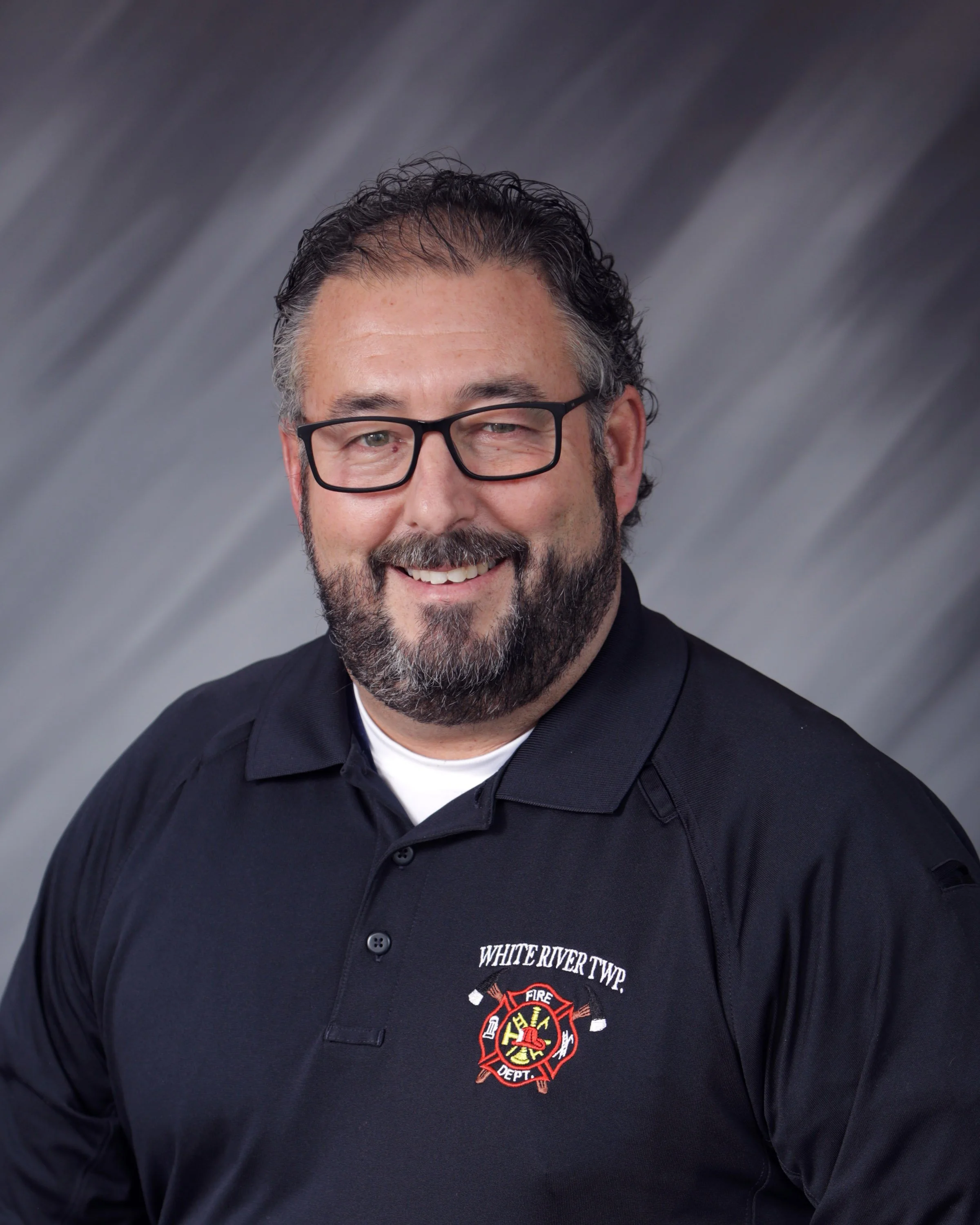 A man with glasses, a beard, and gray hair smiling, wearing a black polo shirt with a White River Township Fire Department logo.