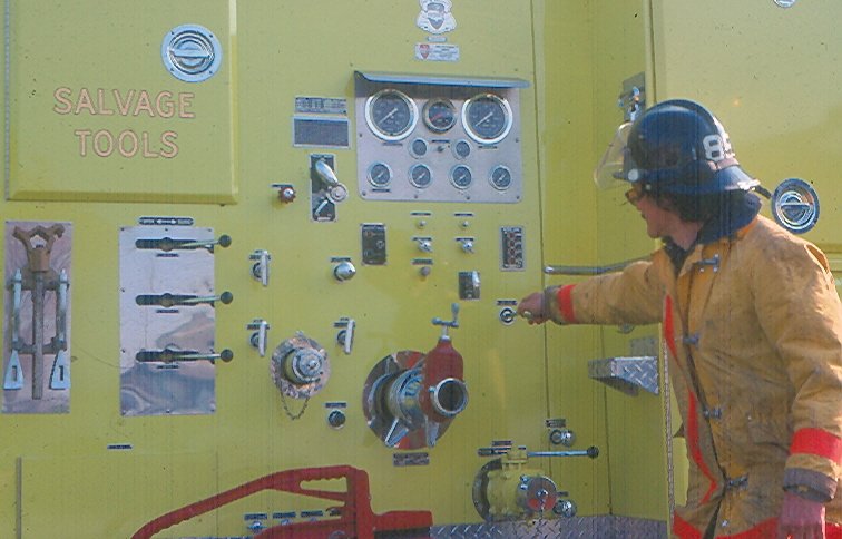 Firefighter in protective gear operating valve on bright yellow fire truck panel with gauges, dials, and salvage tools.