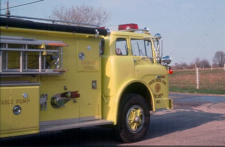 Yellow fire truck parked outdoors during daytime with tools and equipment visible on the side, and a rural background.