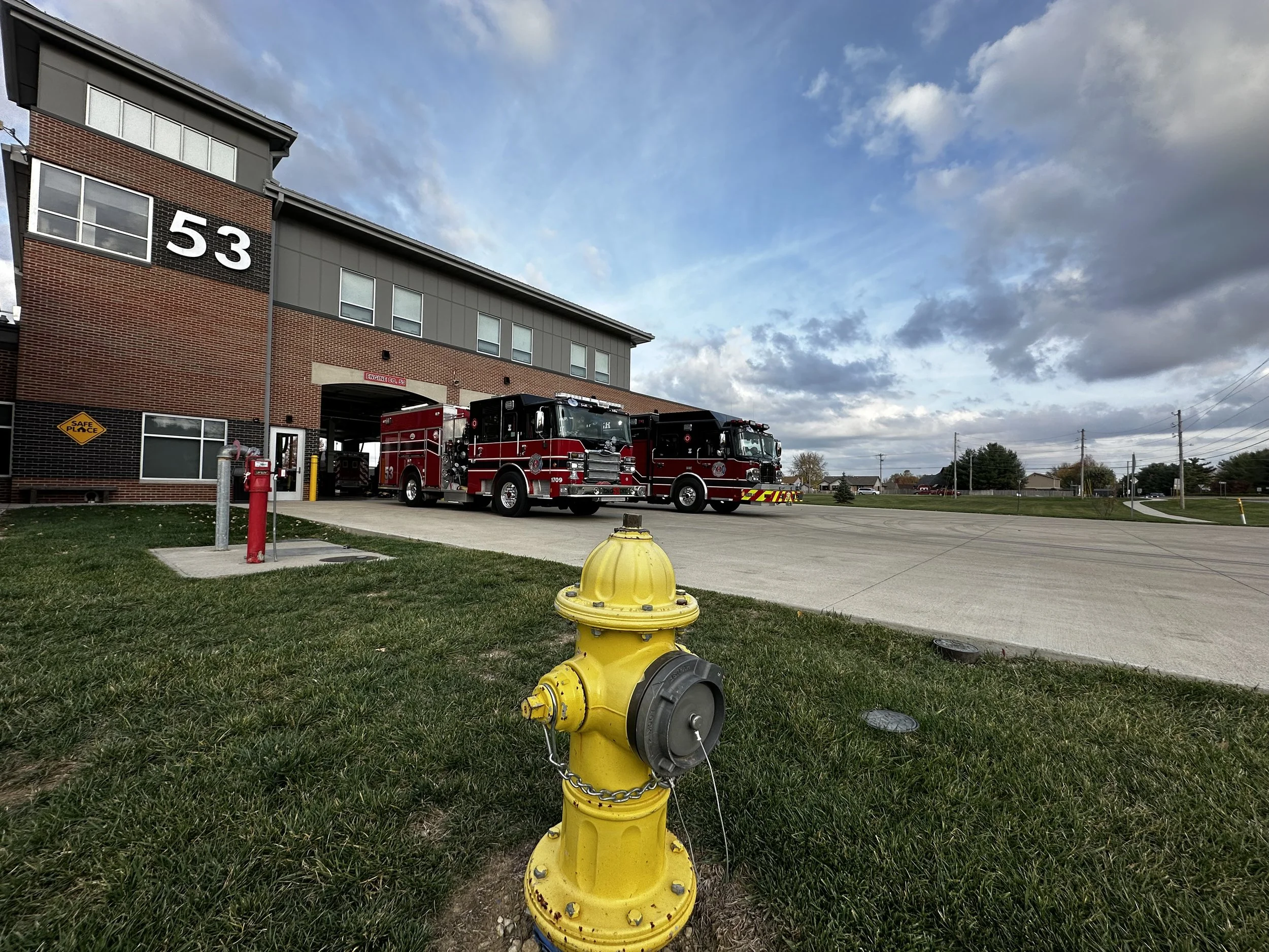 Fire station with two fire trucks parked outside, red building with large number 53, yellow fire hydrant in foreground, cloudy sky.