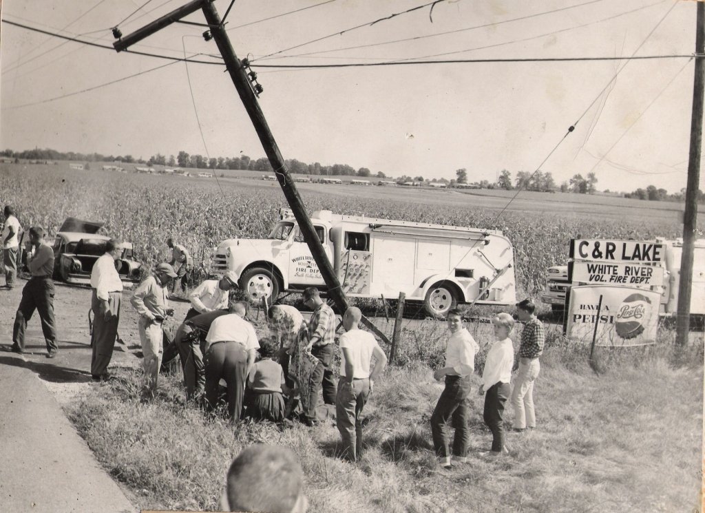 A black-and-white photo of a car accident scene near a roadside. A tilted utility pole is leaning onto a large fire truck in a field. Several people, including men and children, are gathered around inspecting the fallen pole. There are cars parked al