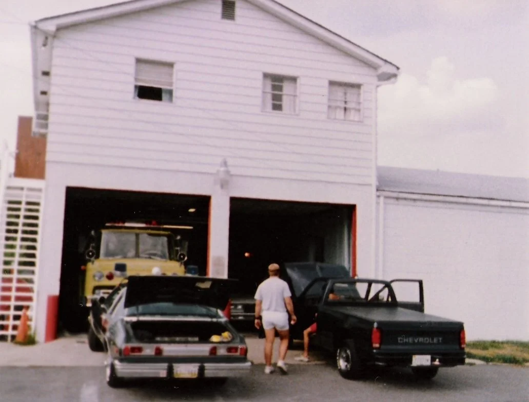 People repairing or working on two cars in front of a white garage with a yellow fire truck inside.