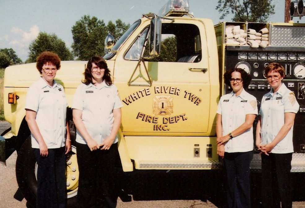 Four women in firefighter uniforms standing in front of a yellow fire truck with the text 'White River Twp Fire Dept Inc.' on the side.