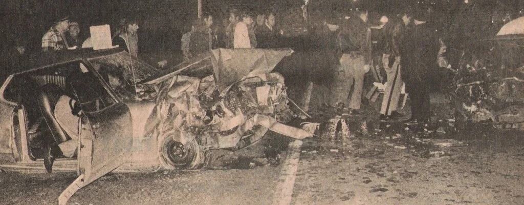 Black and white photo showing a car crash with a seriously damaged vehicle and several people gathered around at night.