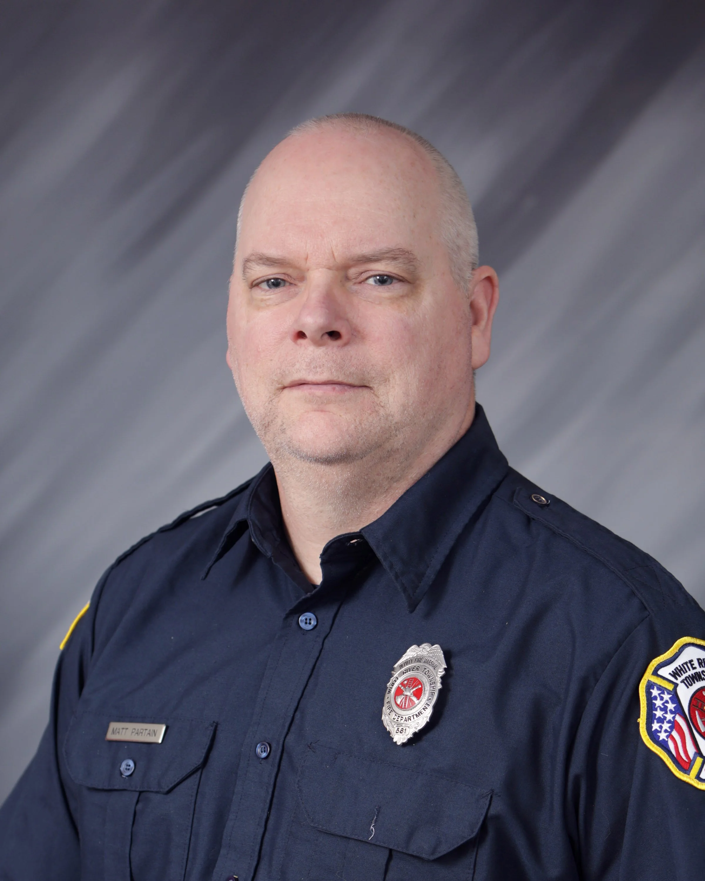 Portrait of a male firefighter in uniform with a badge on his chest and an embroidered patch on his sleeve, against a gray gradient background.