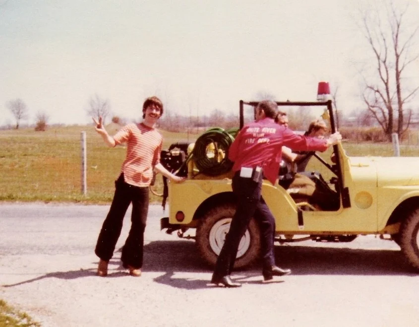 A woman is standing next to a yellow fire truck, smiling and waving, while a firefighter is engaged with the truck on a rural road, with a grassy field and trees in the background.
