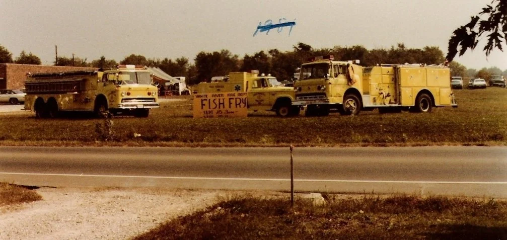 Two yellow fire trucks parked on a grassy area near the road, with a sign advertising fish fry. There are a few cars in the background and trees.
