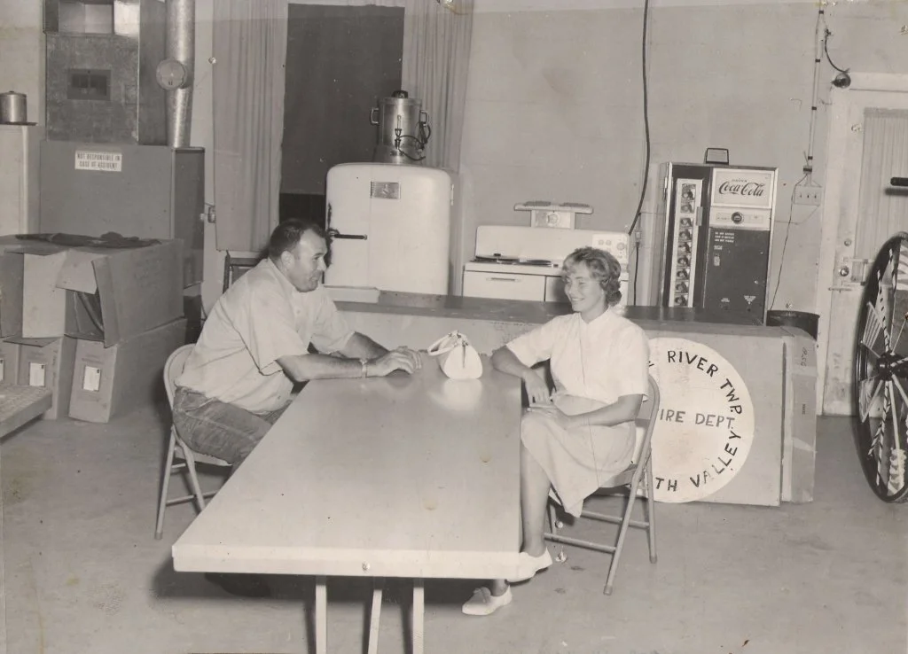 Two people sitting at a long table in what appears to be a kitchen or break room. The man on the left has a beard and is wearing a light-colored shirt, while the woman on the right has short hair and is wearing a dress or uniform. There is a vintage 