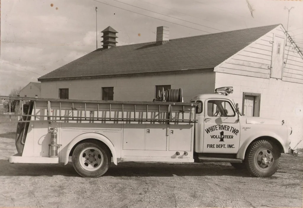 A vintage fire truck with the words "White River Twp Volunteer Fire Dept. Inc." on the side, parked in front of a house with a pitched roof and chimney, in black and white.