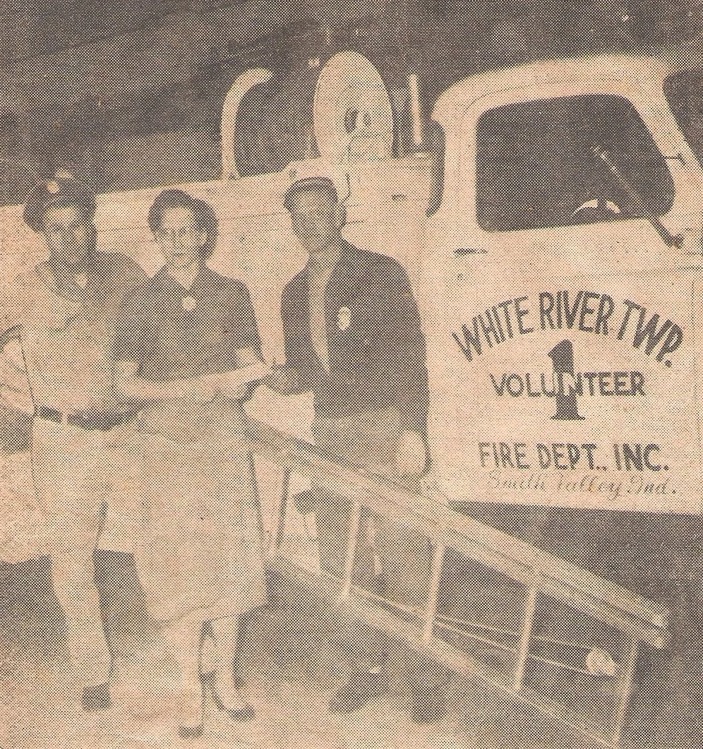 Three individuals standing outdoors near a fire truck marked 'White River Twp. Volunteer Fire Dept. Inc.', with one person handing a plaque to another.
