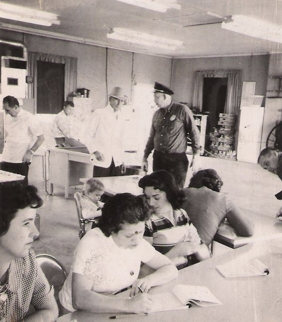 A black-and-white photo of a group of women and children seated at tables in a cafeteria or community center, with three men in uniform standing and walking in the background, possibly staff or officials.