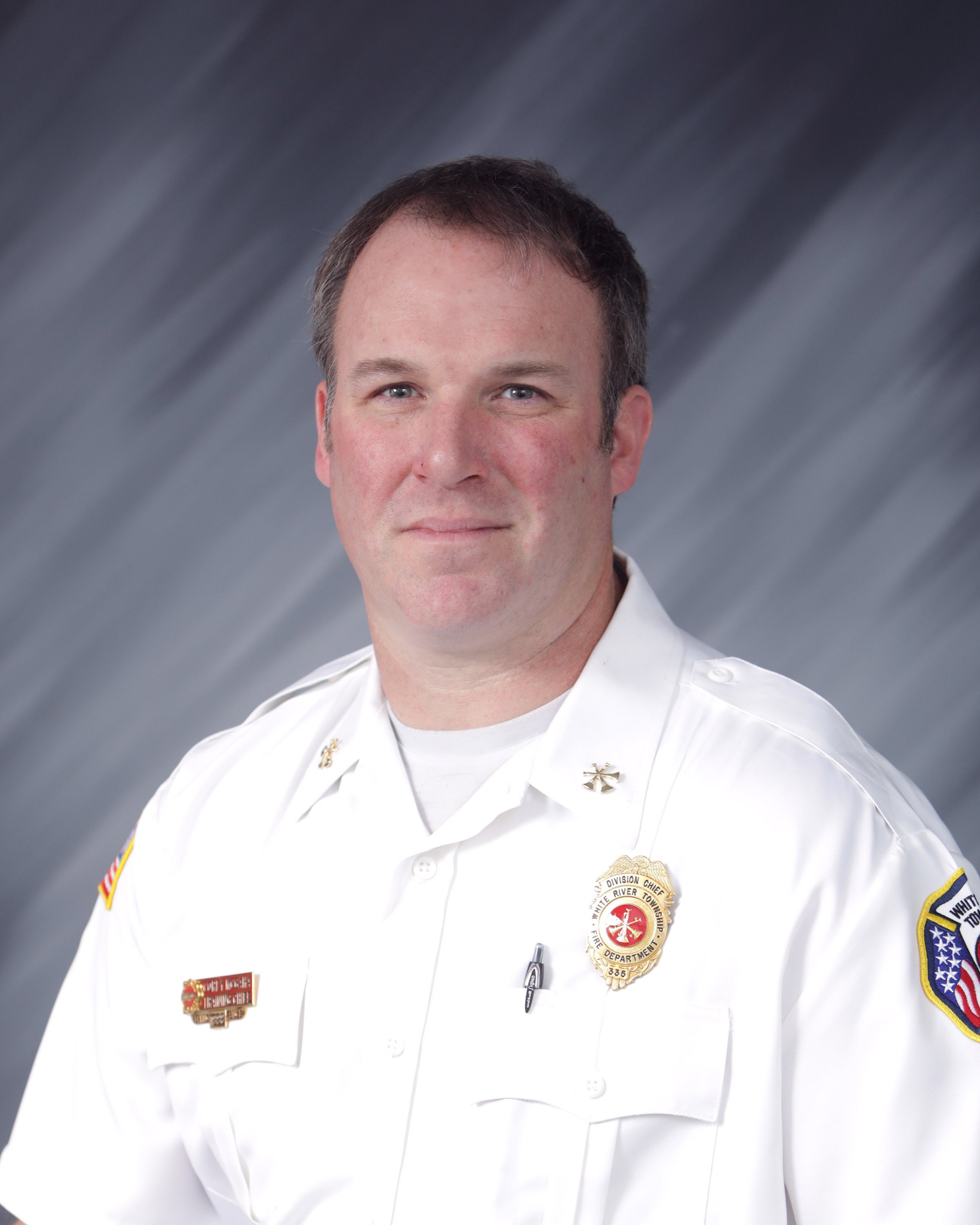 Portrait of a man in a white uniform with patches and badges, against a gray gradient background.