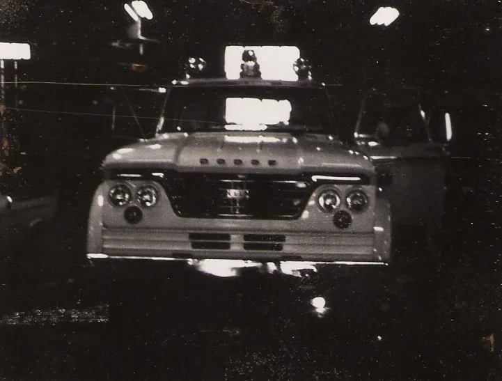 Black and white photo of a vintage race car on a dark track, with bright lights and an audience or officials in the background.
