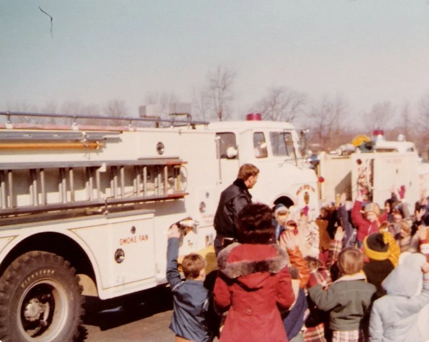 Children and adults gathered around fire trucks during a parade or event on a clear day.