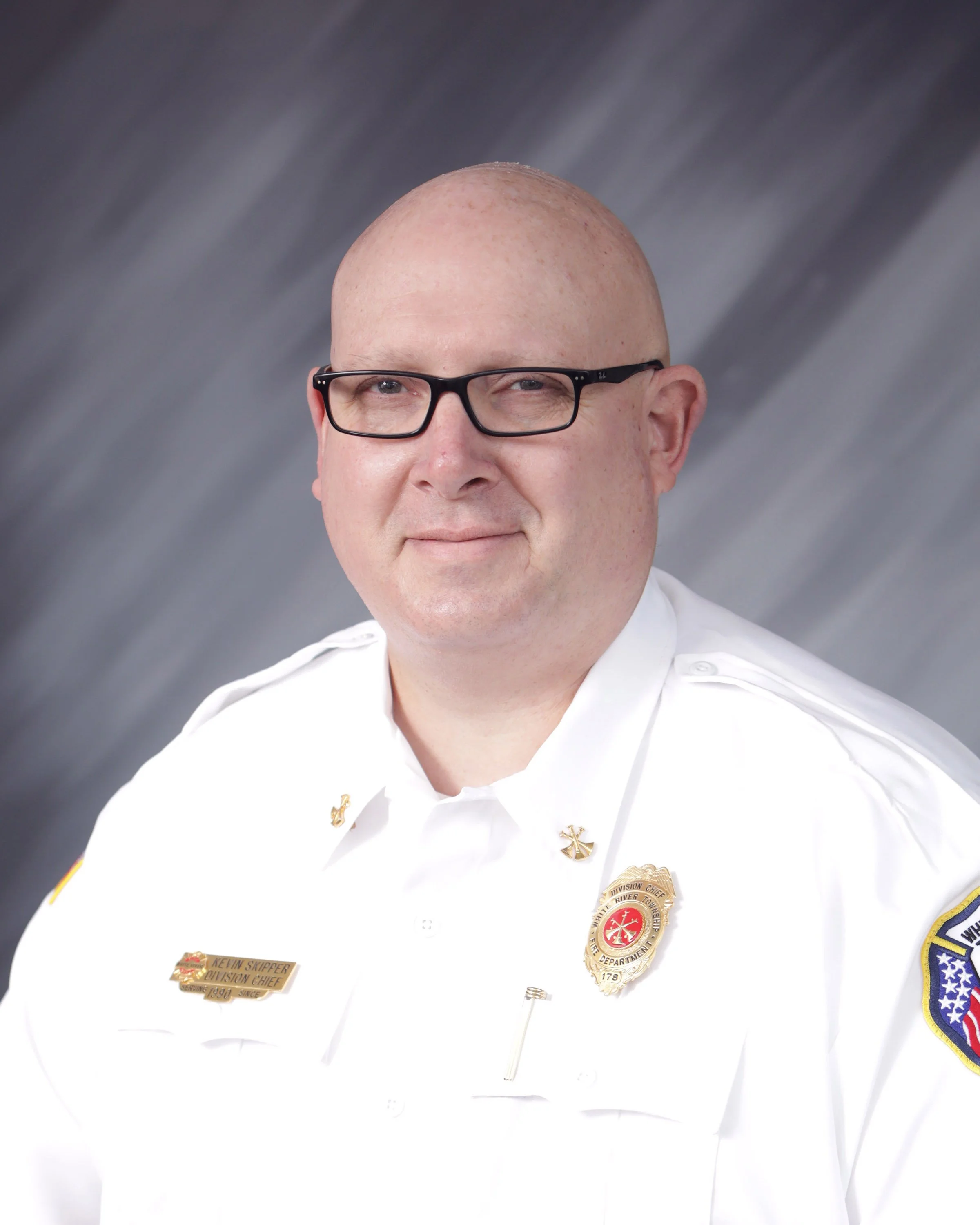 A man in a white firefighter uniform with badges and a patch on the sleeve, wearing glasses, against a gray background.