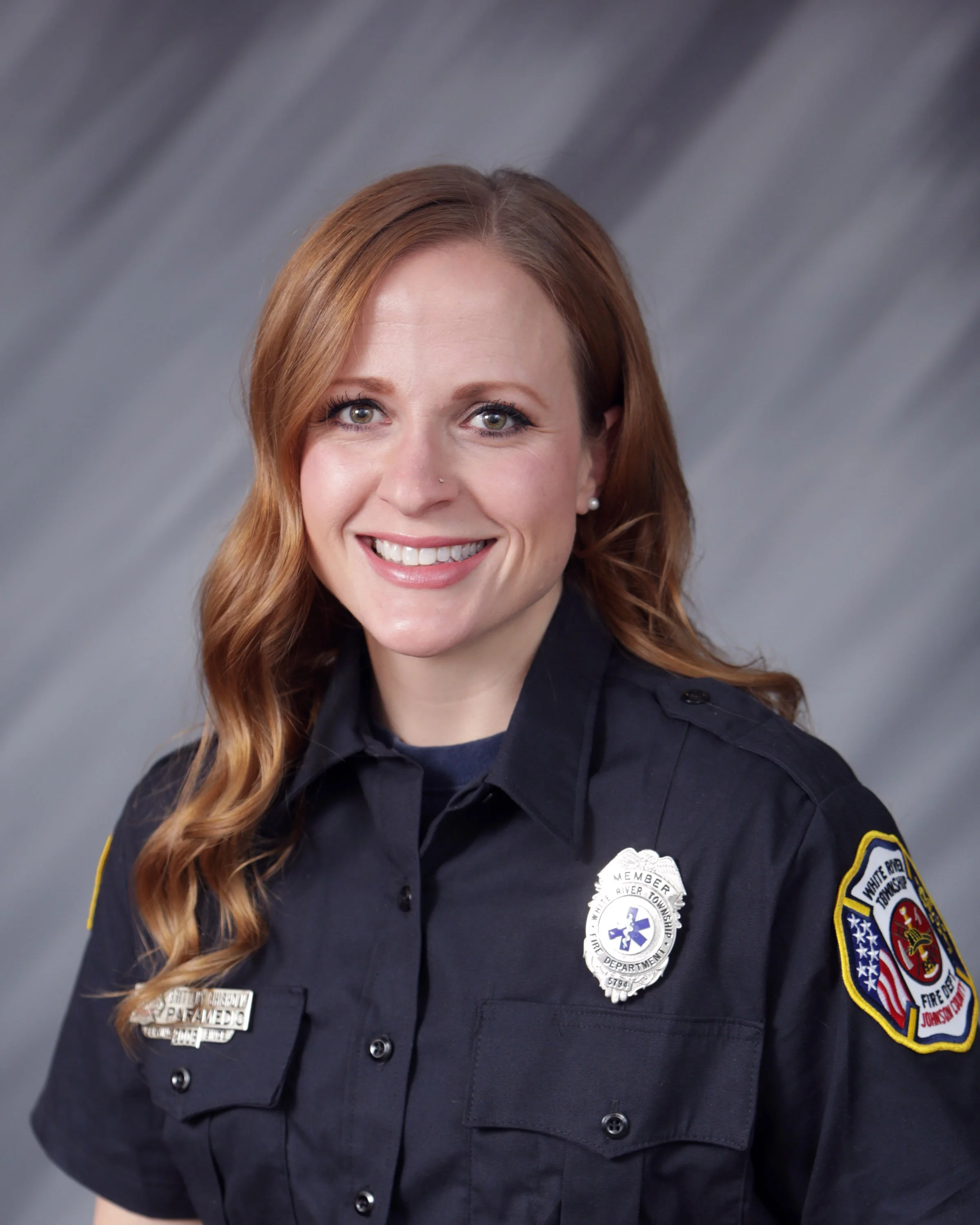 Female firefighter in uniform smiling, with badges and patches on her shirt, gray background.