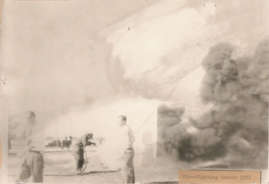 Black and white photo of a fire-fighting training session from 1958, showing firefighters handling hoses with smoke and fire in the background, and a small label at the bottom indicating 'Fire-fighting School 1958'.
