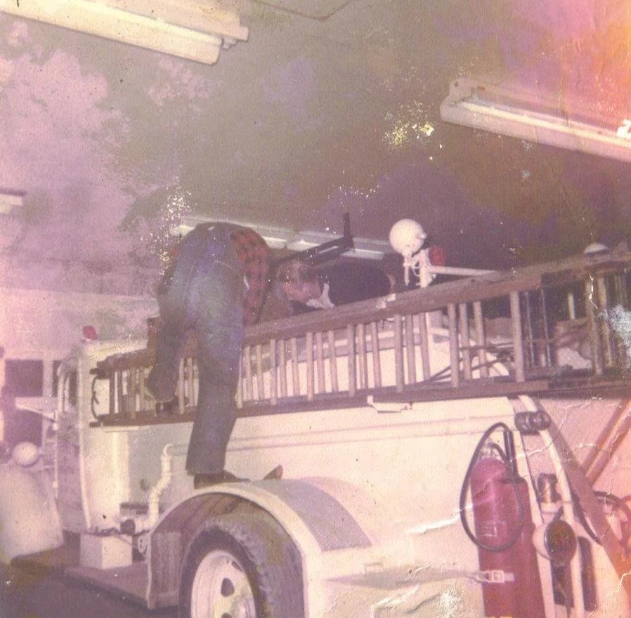 Fire truck parked indoors with a person climbing the ladder and a skeleton statue on top.