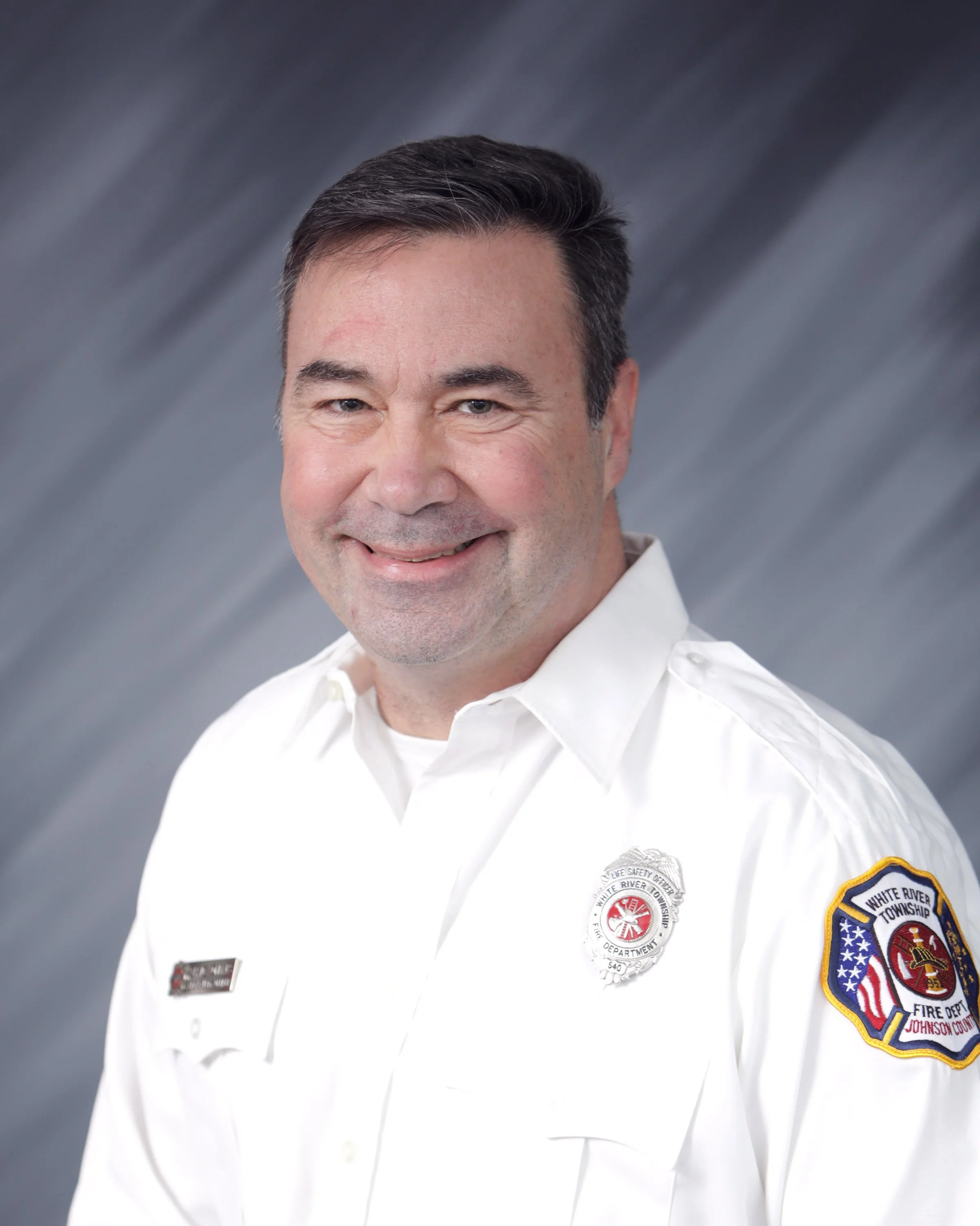 A man in a white uniform with patches and badges, smiling in front of a gray, blurred background.
