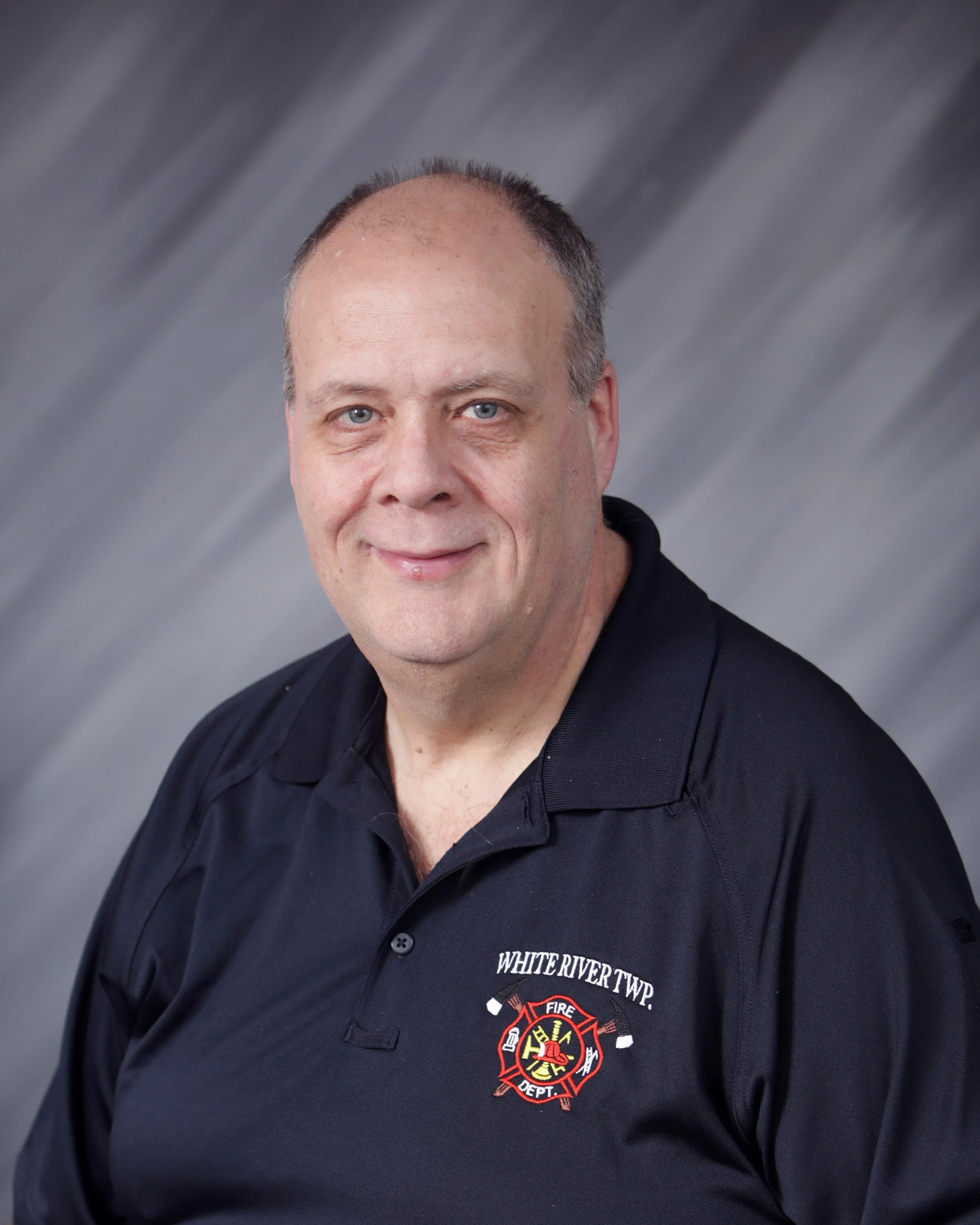 A man with short hair and a friendly expression wearing a black polo shirt with the logo 'WHITE RIVER TWP FIRE DEPT.' on it, against a gray background.
