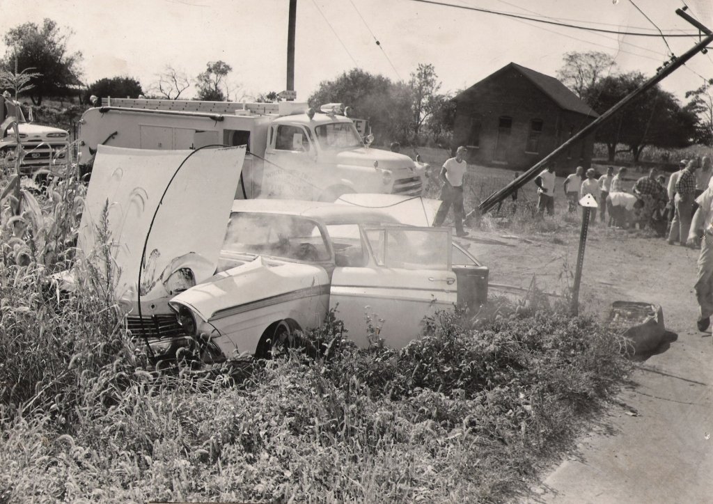 A vintage car crash scene with a car on fire, surrounded by emergency vehicles and people observing, in a rural area.