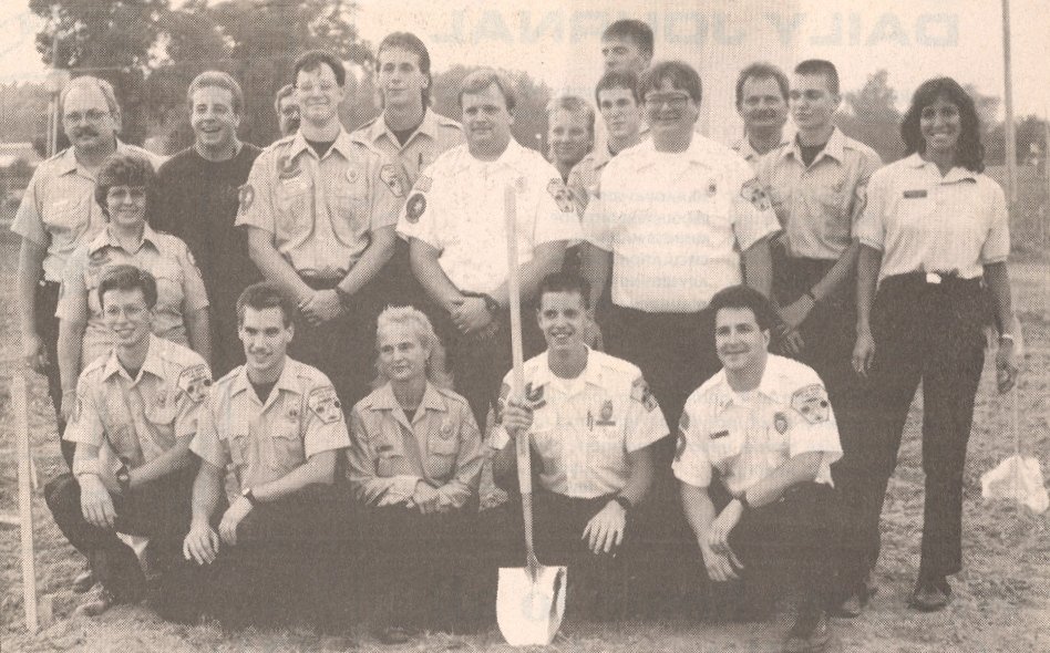 Group of Boy Scouts and adult leaders posing outdoors on a field, some in uniform, one holding a flag, during daytime.