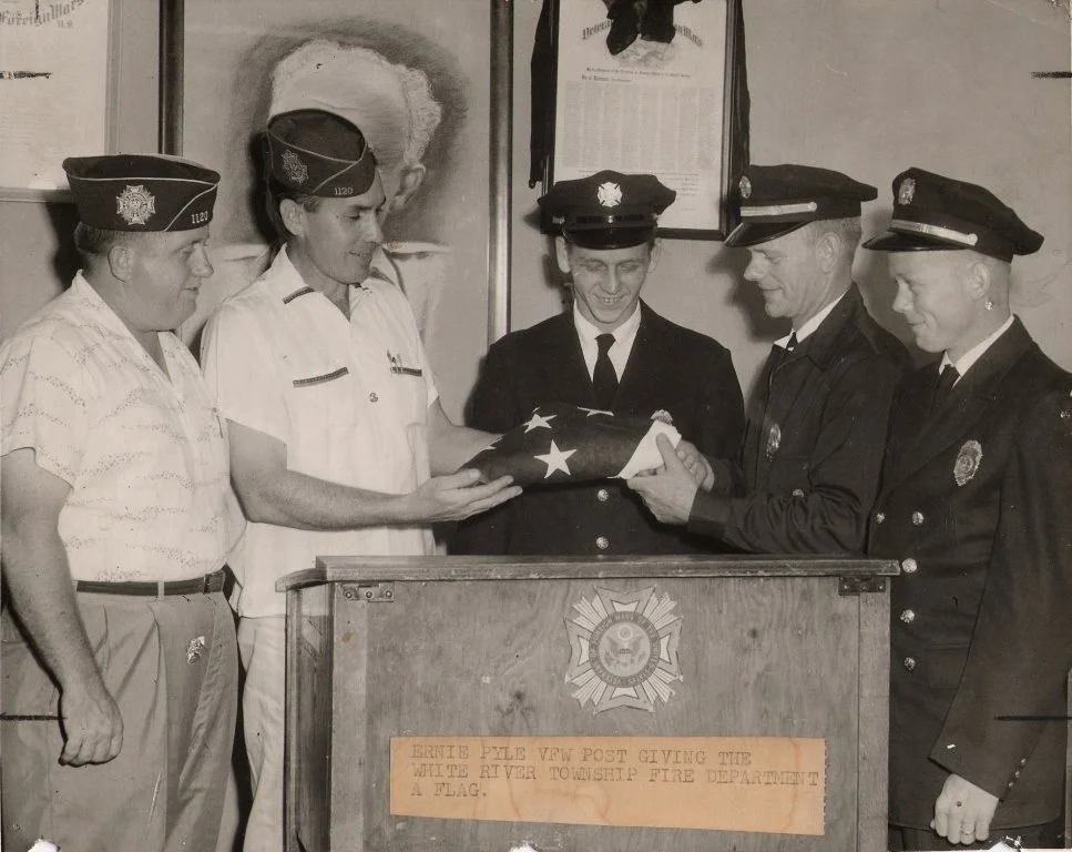 A group of five firefighter and veteran officers are gathered around a wooden podium, presenting or receiving a folded American flag. The background features framed pictures and a plaque, and the officers are dressed in uniforms, some with hats. The 