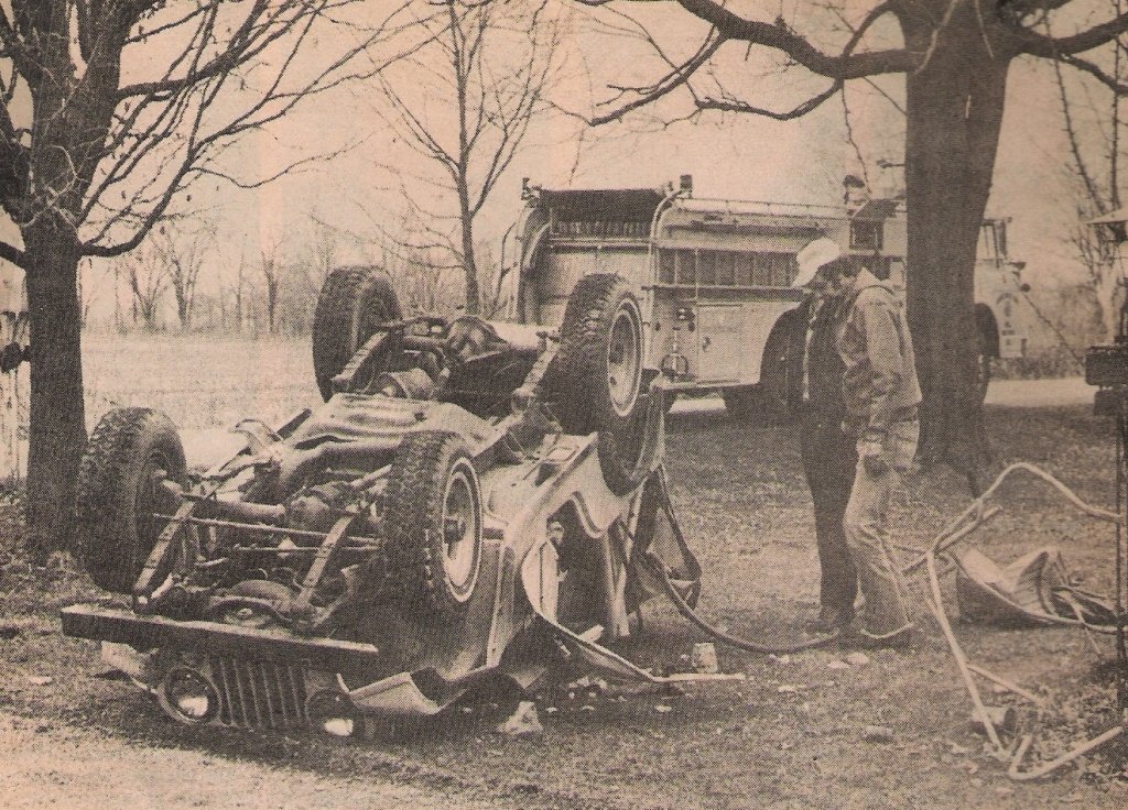 An overturned race car on the grass with a fire truck in the background, trees without leaves, and two men standing nearby, one wearing a cap and the other in a jacket.