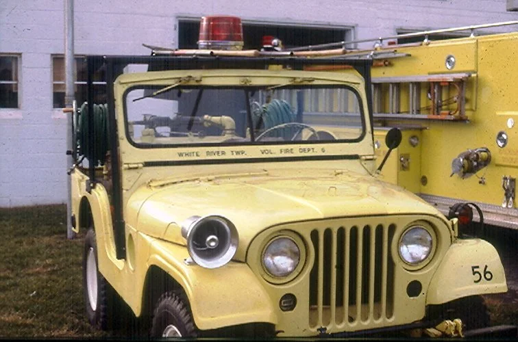 Yellow vintage fire truck with emergency lights and equipment, labeled 'White River Twp. Vol. Fire Dept 6', parked next to a building.