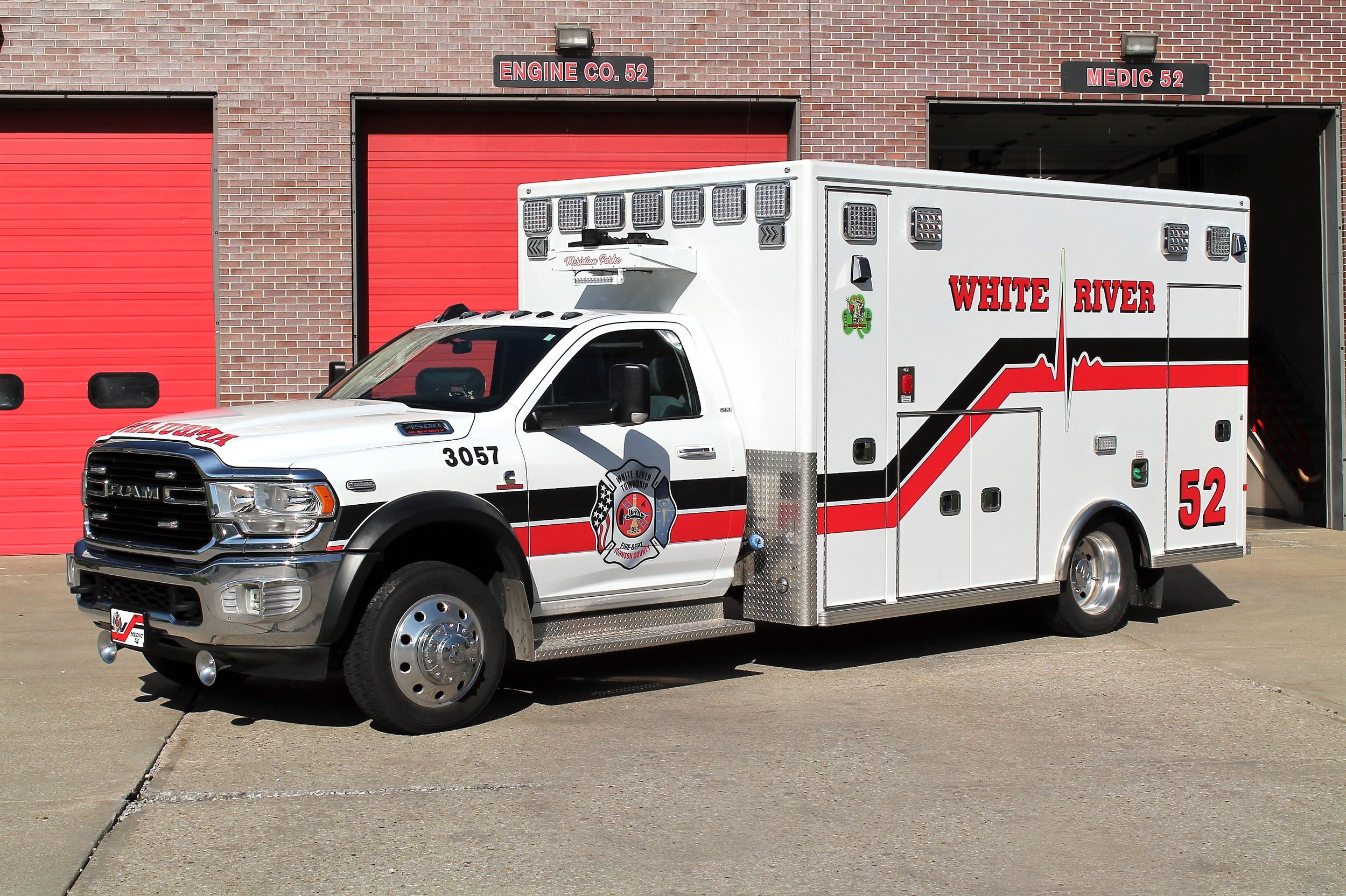 A white emergency vehicle with red and black markings is parked outside a fire station. The vehicle has the words 'White River' and the number 52 on its side, along with a fire department emblem. The garage behind it has two red doors and signs reading 'Engine Co. 52' and 'Medic 52'.