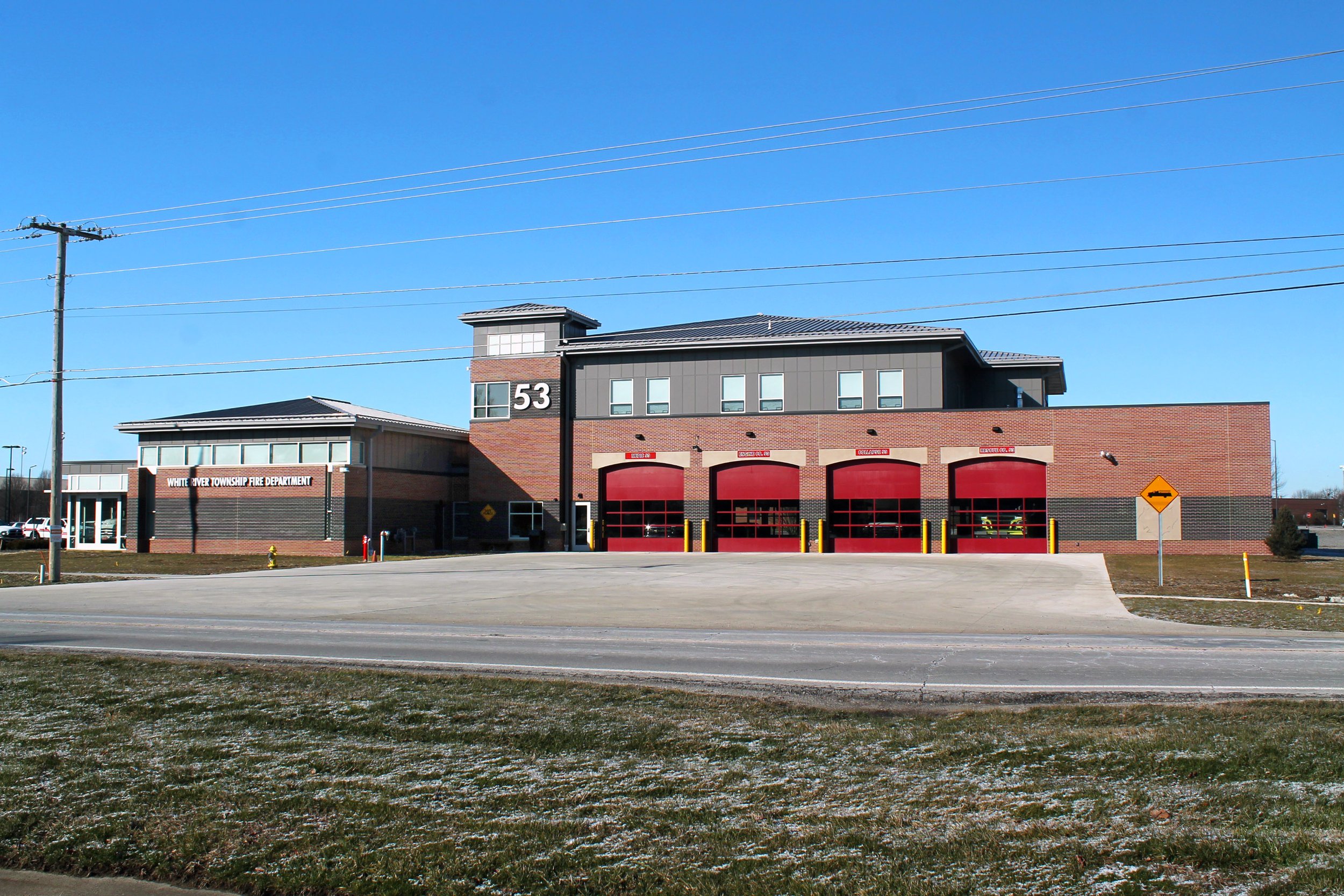 Front view of a modern fire station with three red garage doors, a black sign with white letterings, and the number 53 on the building, under a clear blue sky.