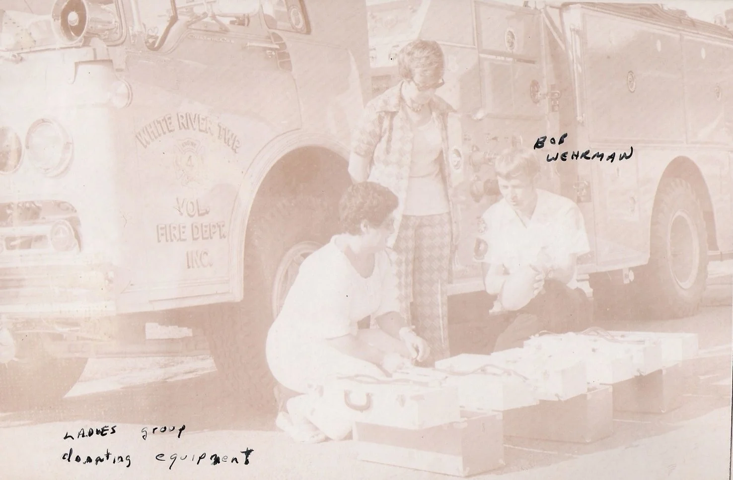A vintage black and white photo of three women kneeling by a collection of boxes in front of a fire truck with the words 'White River Township Volunteer Fire Department Inc' visible on it. The women are identified as members of the Ladies Group donat