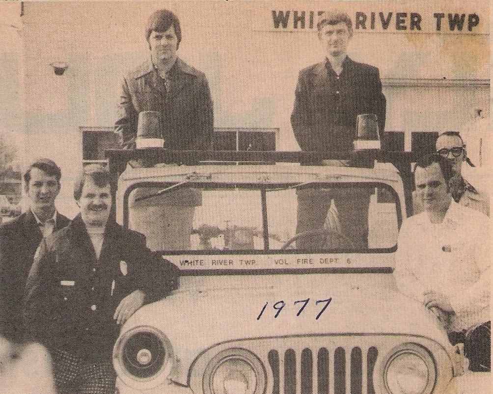 Group of people posing around a fire truck in front of a building with a sign that reads 'White River Twp,' dated 1977.