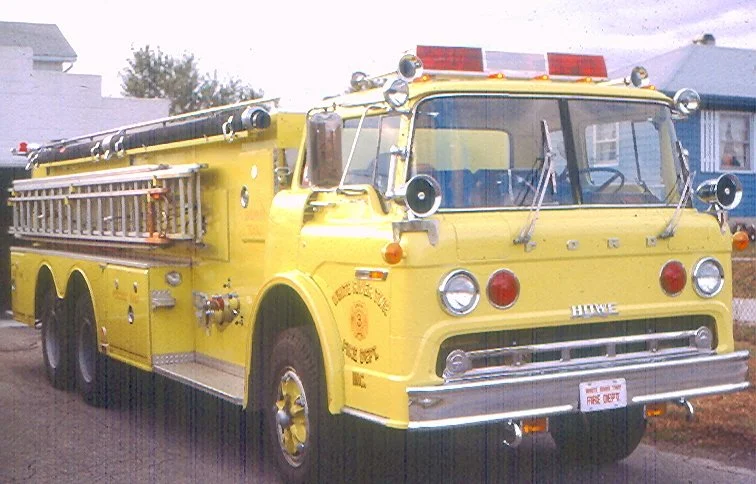 Yellow vintage fire truck with ladder, sirens, and emergency lights parked on a residential street.