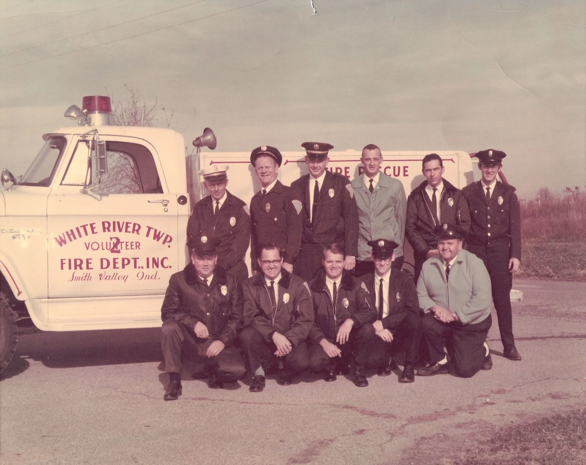 A group of eleven firefighters and volunteers posing in front of a vintage White River Township fire truck. The group includes both men and women dressed in firefighter uniforms and casual jackets.
