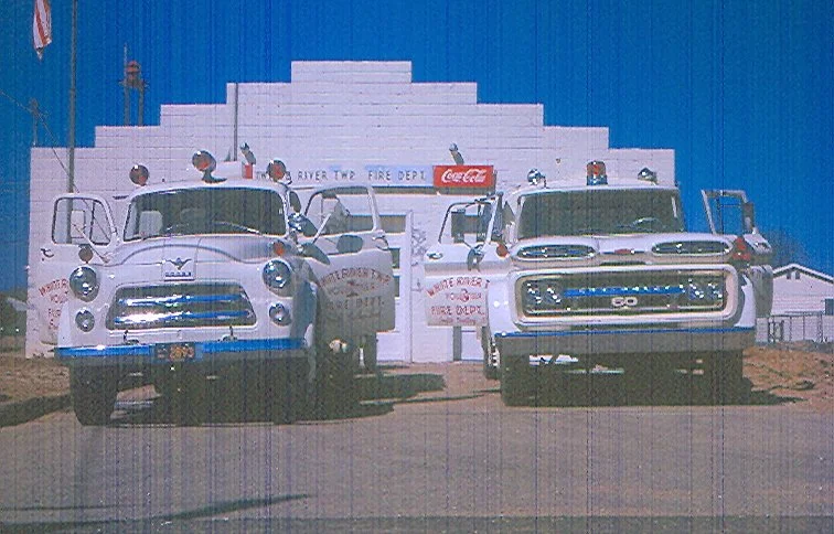 Two vintage fire trucks parked in front of a building with a blue facade, with signs indicating a fire department and Coca-Cola advertisement.