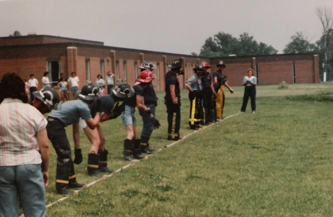 Group of firefighters in firefighting gear practicing on a field with a rope line, with onlookers in the background.