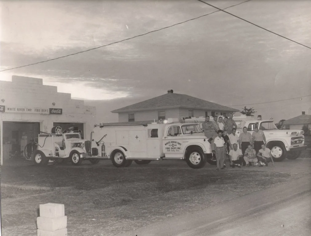 Black and white photo of the White River Township Fire Department with fire trucks and firefighters, a group of people standing and squatting in front of the vehicles, houses and an overcast sky in the background.