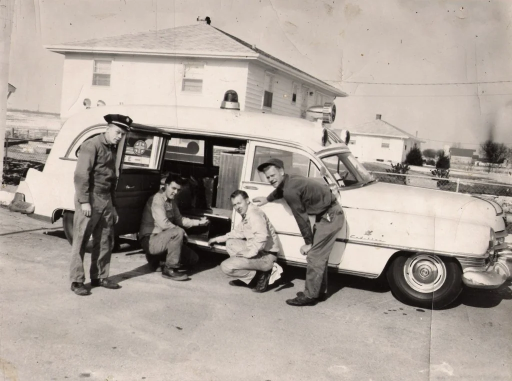 Four men in uniform gather around an ambulance vehicle with a house in the background.